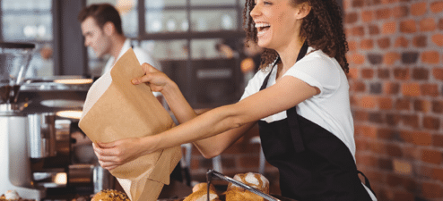 A cheerful barista handing a paper bag filled with baked goods to a customer in a cozy bakery, illustrating customer engagement strategies to increase foot traffic and customer satisfaction.